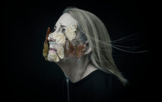 A woman against a black background looks in to the distance wistfully. Her face is covered in butterflies, which add a touch of color to the photo and all but cover a breathing tube on the woman's face.